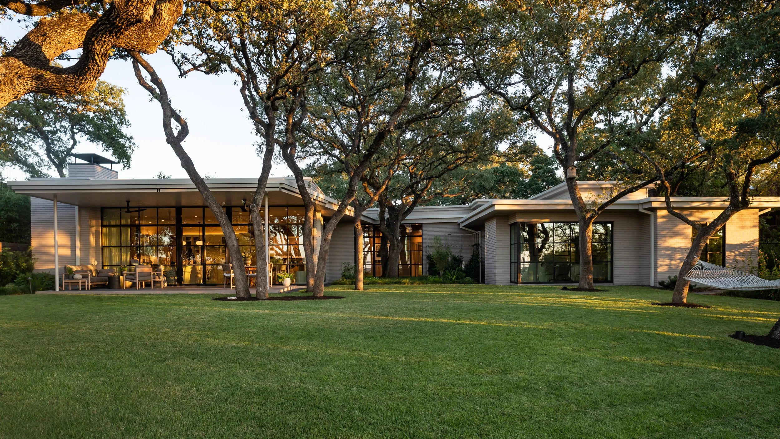 Back exterior view of Austin mid-century modern home with covered outdoor sitting and dining areas surrounded by mature oak trees.