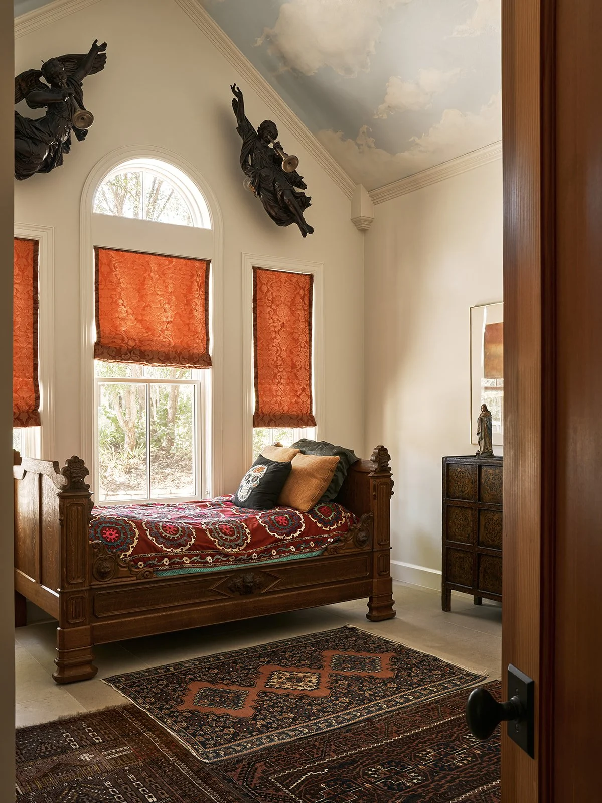 “Cloud room” guest bedroom in an Austin interior design project featuring hand-painted cloud ceiling murals, a vintage wood daybed, layered antique rugs, silk brocade window shades, carved angel sculptures, and a Tibetan chest.