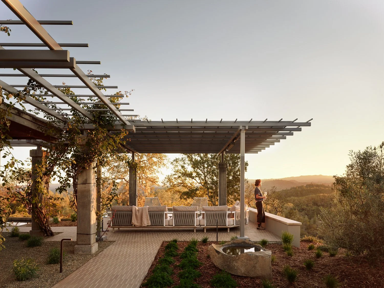 Roda outdoor lounge seating beneath an architectural trellis, framed by rolling wine country hills, with sculptural stone elements and warm evening light creating a relaxed outdoor living space.
