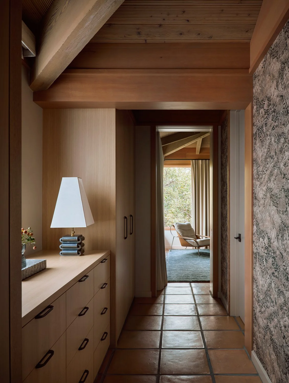 White oak millwork hallway with Jim Thompson wallpaper, custom built-in storage, and a blue cashmere rug softening the original Saltillo tile floors, leading toward a corner window and Zanotta lounge chair overlooking the Sonoma landscape.