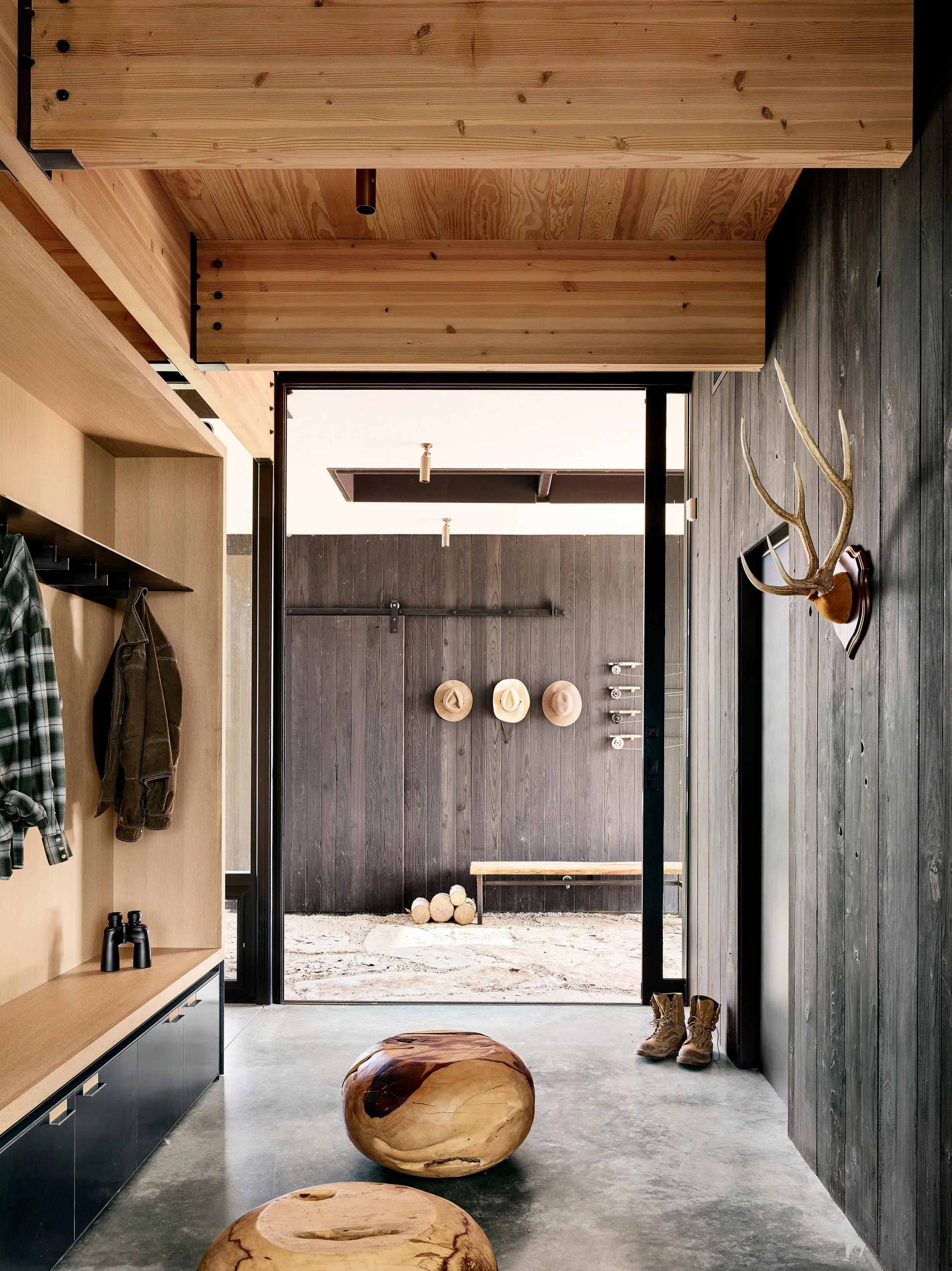 Mudroom with built-in storage, space for boots and jackets, antlers sourced from the property, and solid wood ottomans that bring the outdoors inside, opening to the dogtrot beyond.