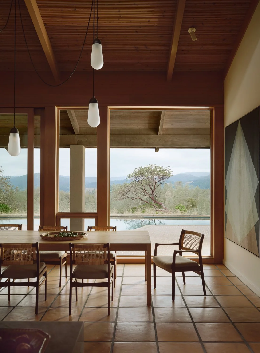 Dining room opening to the pool and wine country landscape, featuring a custom white oak dining table, woven wicker and oak chairs, a Tassel chandelier by Apparatus, and a large painting by Gonzalo Lebrijo framed by expansive wood windows.