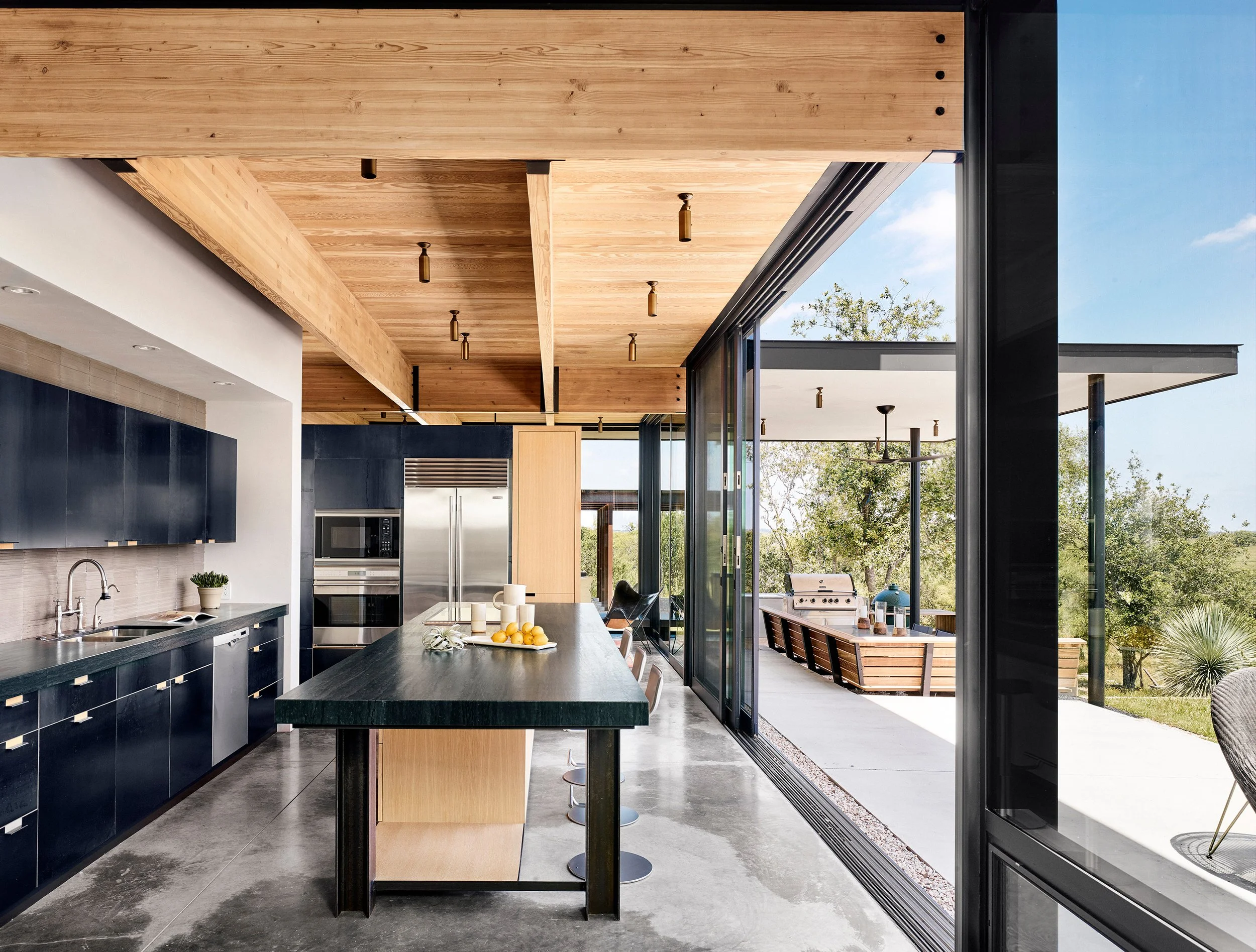 Kitchen in a modern Hill Country home featuring a white oak island, dark stone countertops, steel-paneled cabinetry, concrete floors, and large sliding glass doors opening to the outdoor terrace.