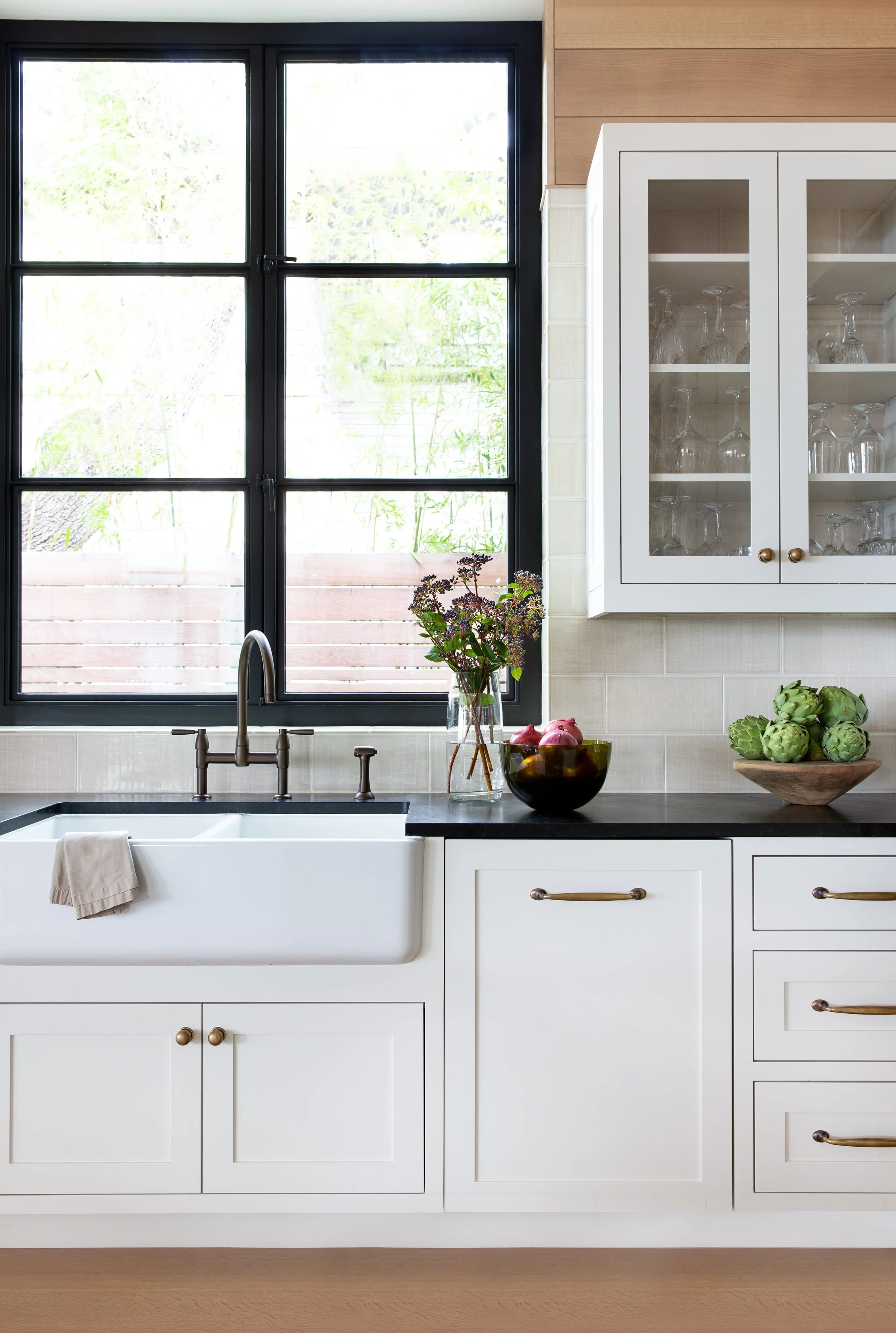 Kitchen sink detail featuring Shaw farmhouse sink, steel window, inset European cabinetry, glass-front cabinets, and antique brass hardware.