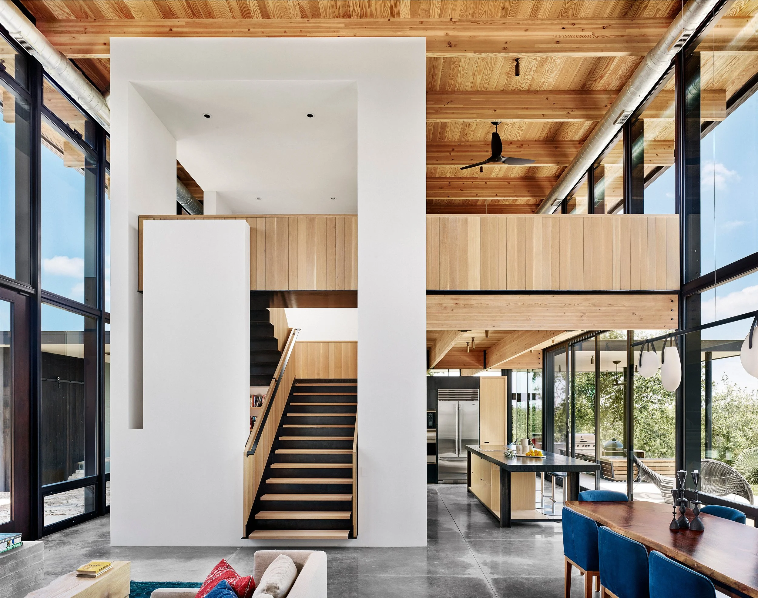 Interior of a modern Hill Country home designed by Laura Roberts Design, showing the kitchen, dining and stairwell to the second floor with plaster walls, concrete floors, white oak millwork, and expansive glazing.