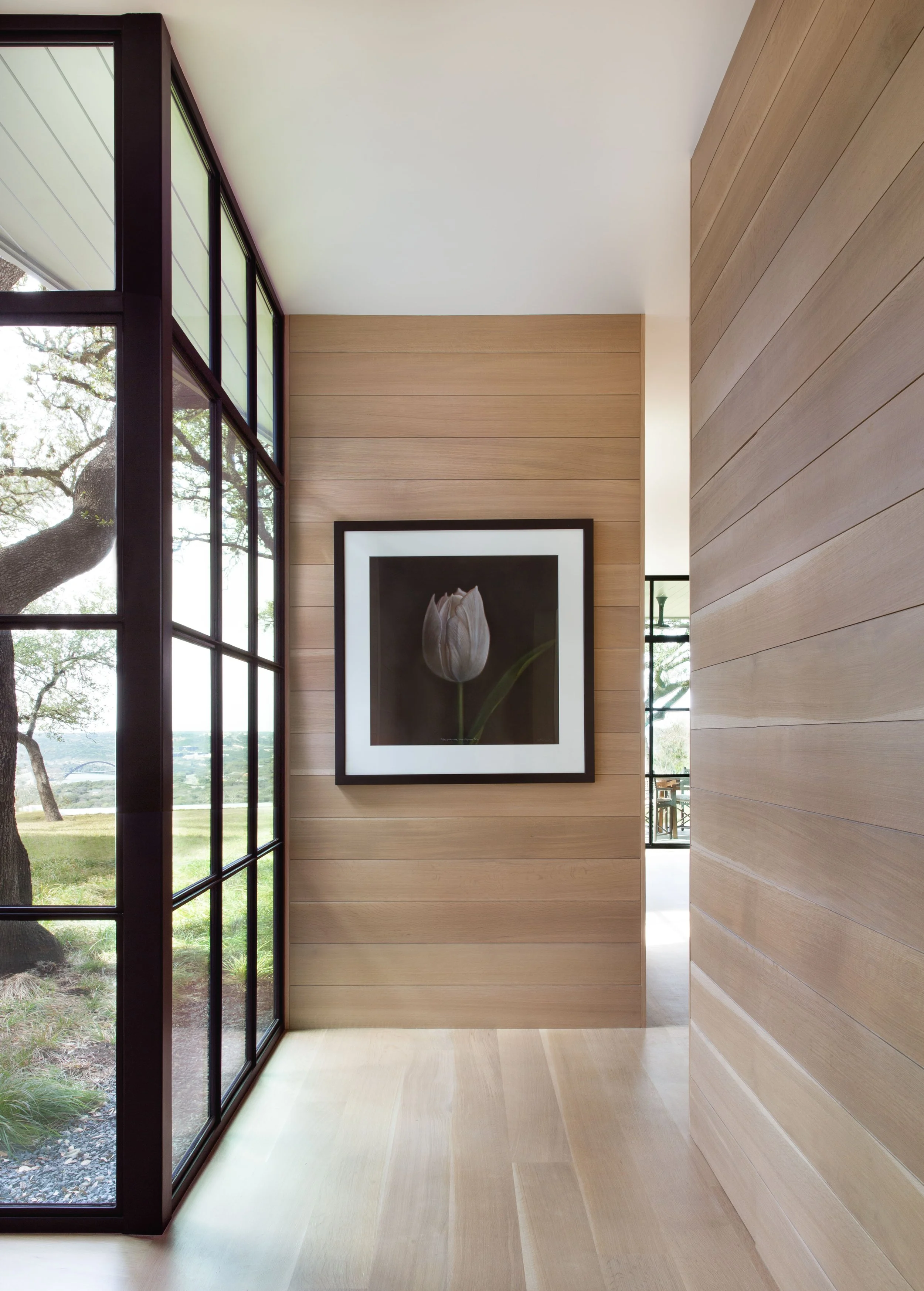 Hallway view with white oak shiplap walls and floors, floor-to-ceiling steel window, and Kate Breakey tulip photograph overlooking the Austin hills.