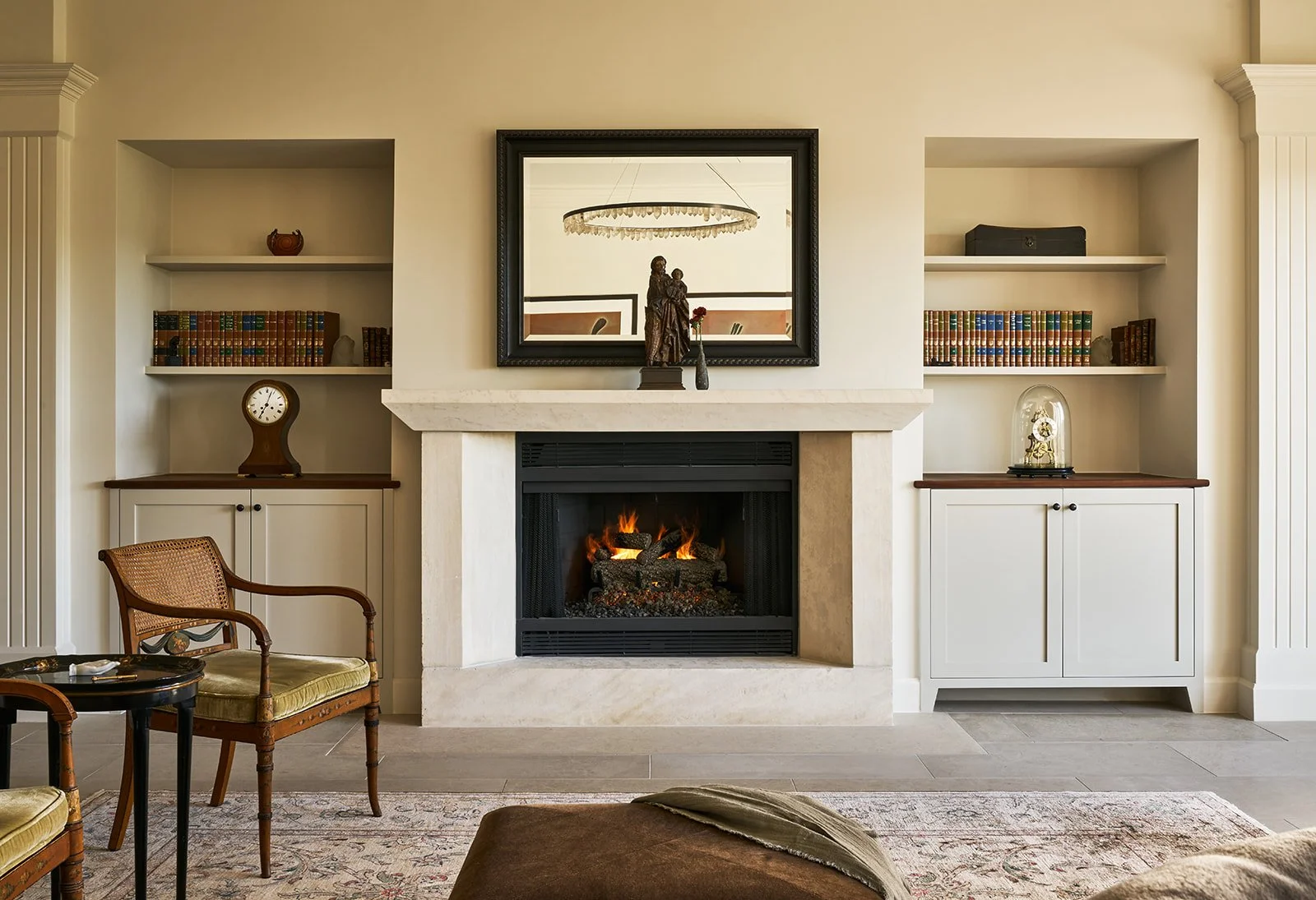 Austin interior design living room by Laura Roberts Design featuring a Christopher Boots Prometheus crystal chandelier, limestone fireplace, Lueders stone floors, vintage chair, clocks, books, and custom built-in bookshelves.