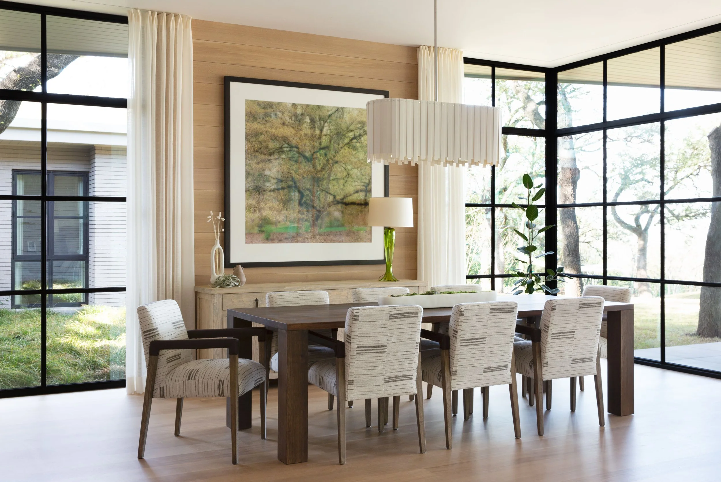 Austin dining room with custom upholstered chairs, large Parsons table, Lynn Geesaman tree photograph, porcelain chandelier, and full-height steel windows.
