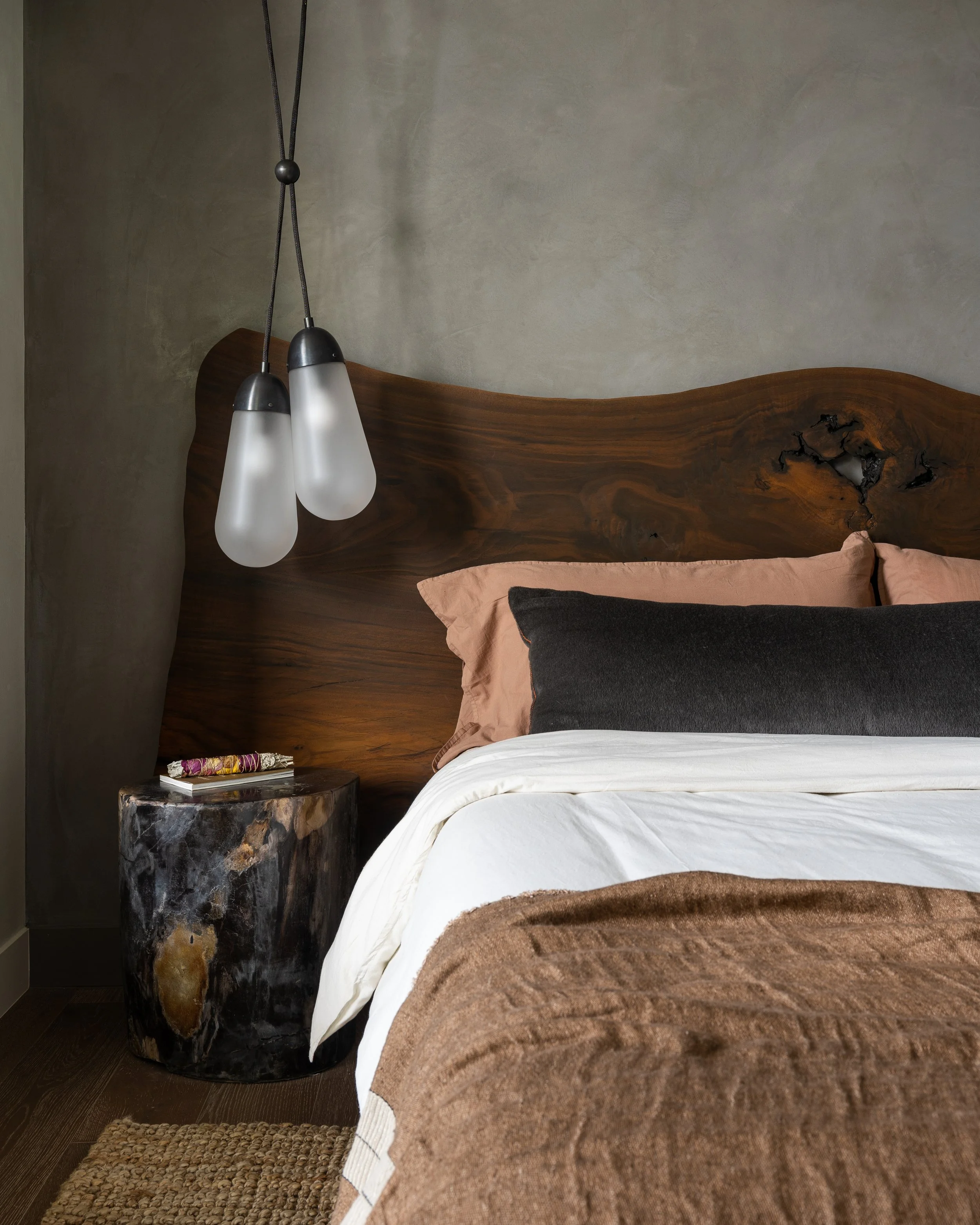 Primary bedroom featuring a custom solid walnut bed crafted from a single live-edge slab, petrified wood side table, cozy neutral bedding, and Apparatus tassel pendants.