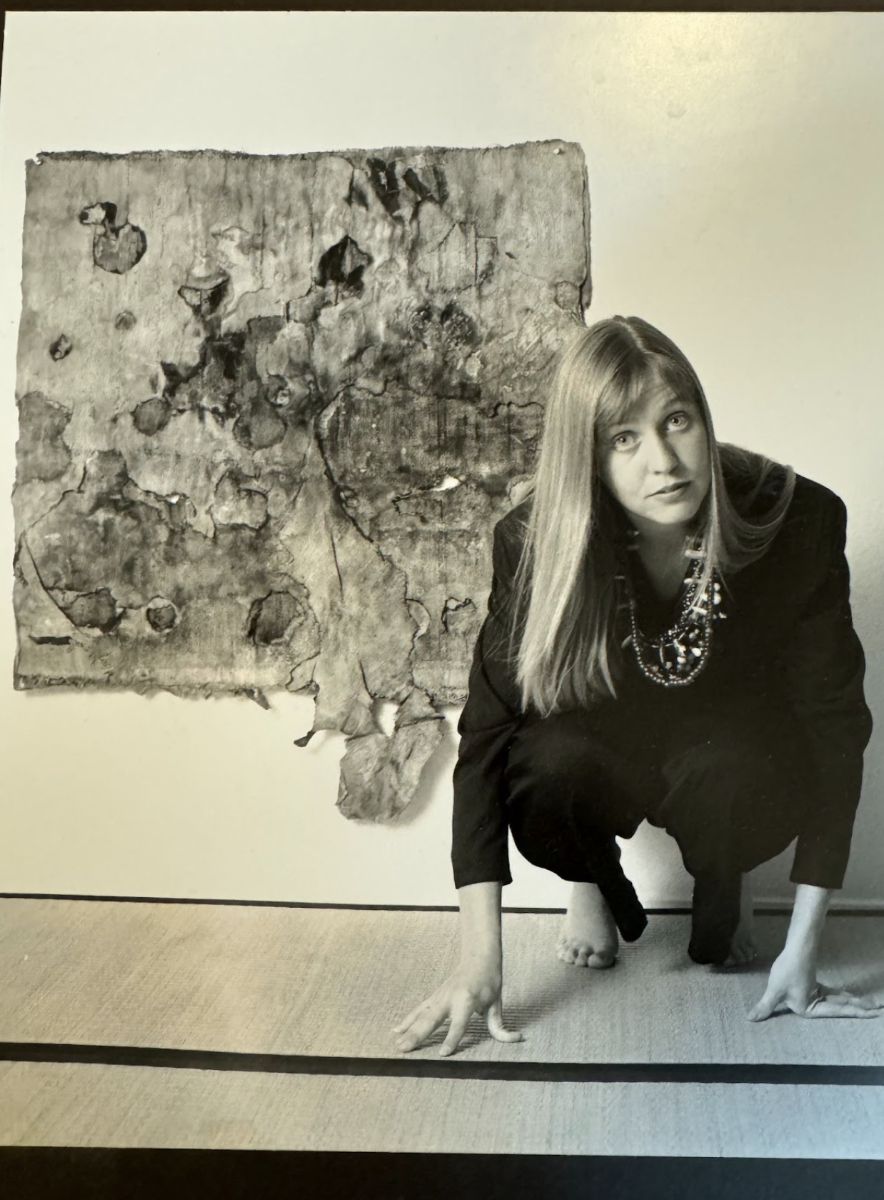 Portrait of Laura Nacke seated on tatami mats beside an early textile weaving artwork, burned and layered back together, in a studio setting